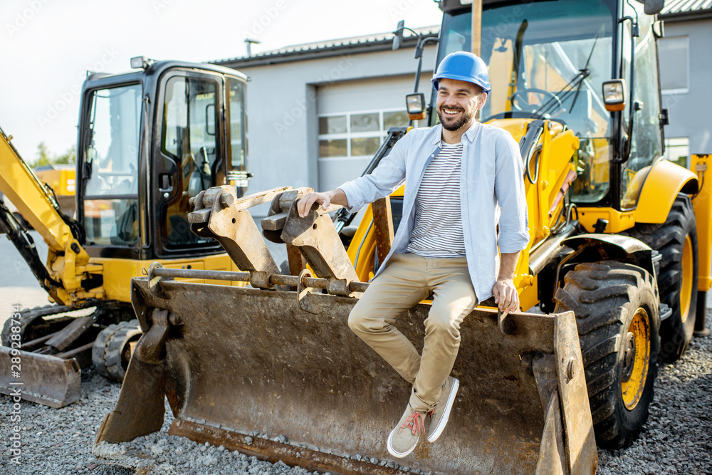 Portrait of a handsome builder sitting on a bulldozer scoop on the open ground of the shop with heavy machinery