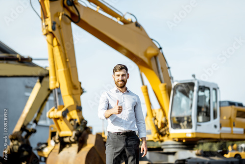 Portrait of a handsome sales consultant or manager standing on the open ground of the shop with heavy machinery for construction