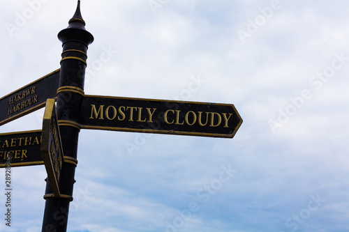 Text sign showing Mostly Cloudy. Business photo text Shadowy Vaporous Foggy Fluffy Nebulous Clouds Skyscape Road sign on the crossroads with blue cloudy sky in the background