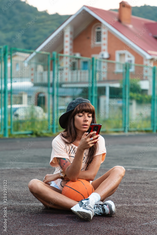 A beautiful young woman with tattoos, wearing a cap, sitting on a sports court with a basketball and looking into a mobile phone