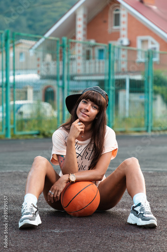 Beautiful young woman with tattoos, wearing a cap, sitting on the sport court with a basketball