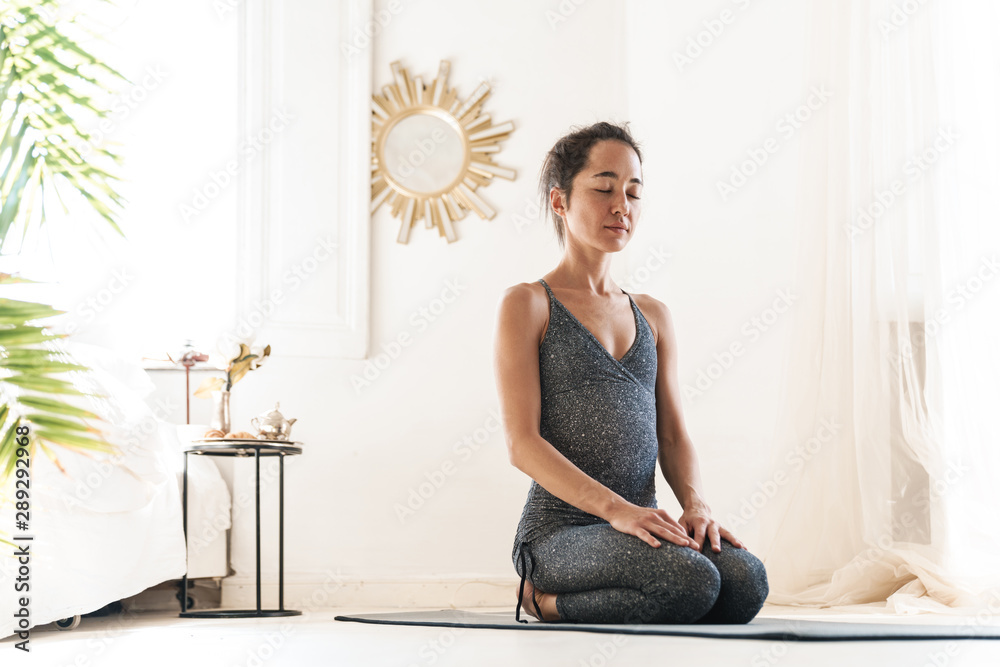 Photo of concentrated sportswoman sitting on fitness mat and meditating while practicing yoga in bright room Photo of concentrated sportswoman sitting on fitness mat and meditating while practicing yoga in bright room