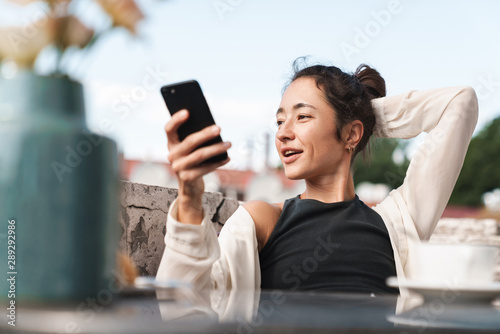 Portrait of happy brunette woman holding smartphone and drinking coffee while resting on terrace outdoors