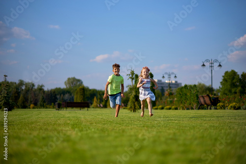 Two happy little kids having fun while running through the grassy field and racing against each other. children running around the green lawn, happy and cheerful, running away to the summer park