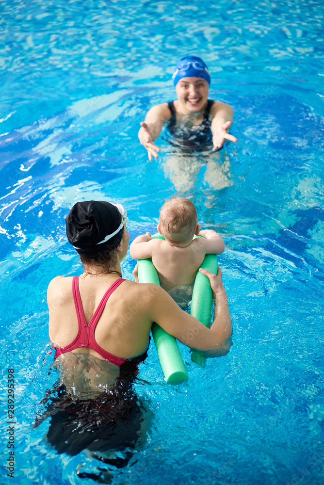 Mother with child in paddling pool. Instructor teaching little child swimming with pool noodles for protection. Back view of baby floating to his mom. Prevention of diseases of musculoskeletal system