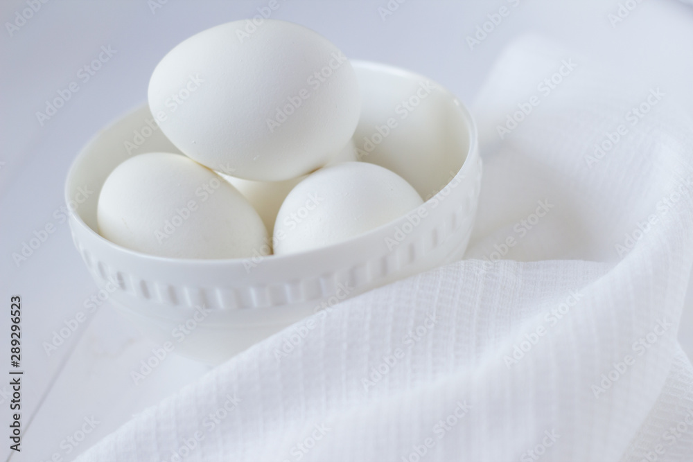 four white eggs lie in a plate on a white wooden table next to a towel. four white eggs lie in a plate on a white wooden table next to a towel.