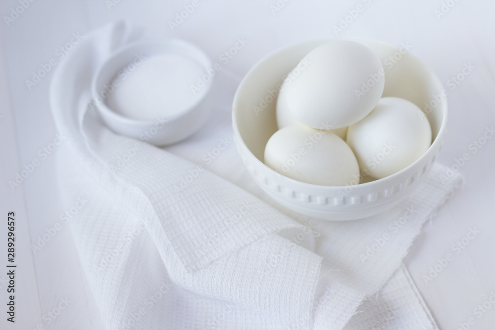 four white eggs lie in a plate on a white wooden table next to a towel and salt shaker. four white eggs lie in a plate on a white wooden table next to a towel and salt shaker.