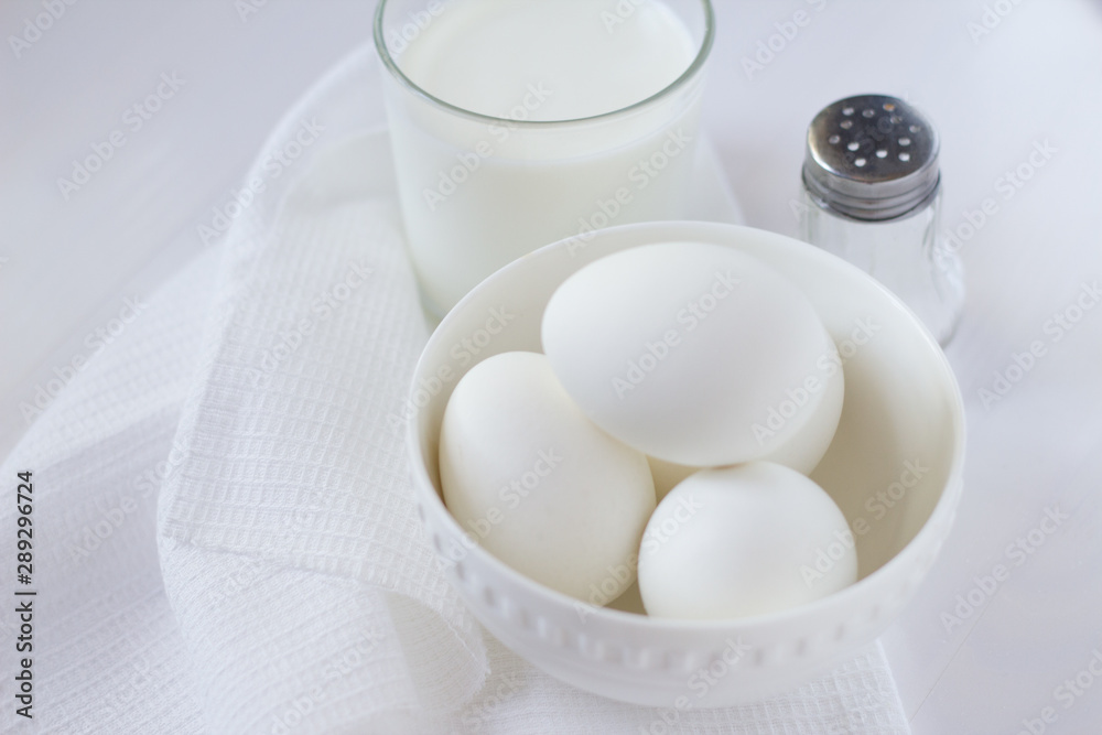 four white eggs lie in a plate on a white wooden table next to a towel and a glass of milk and a salt shaker. four white eggs lie in a plate on a white wooden table next to a towel and a glass of milk and a salt shaker.