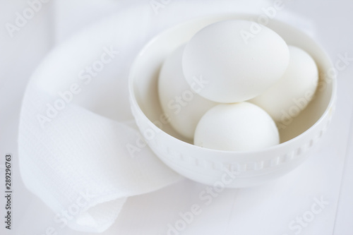 four white eggs lie in a plate on a white wooden table next to a towel.