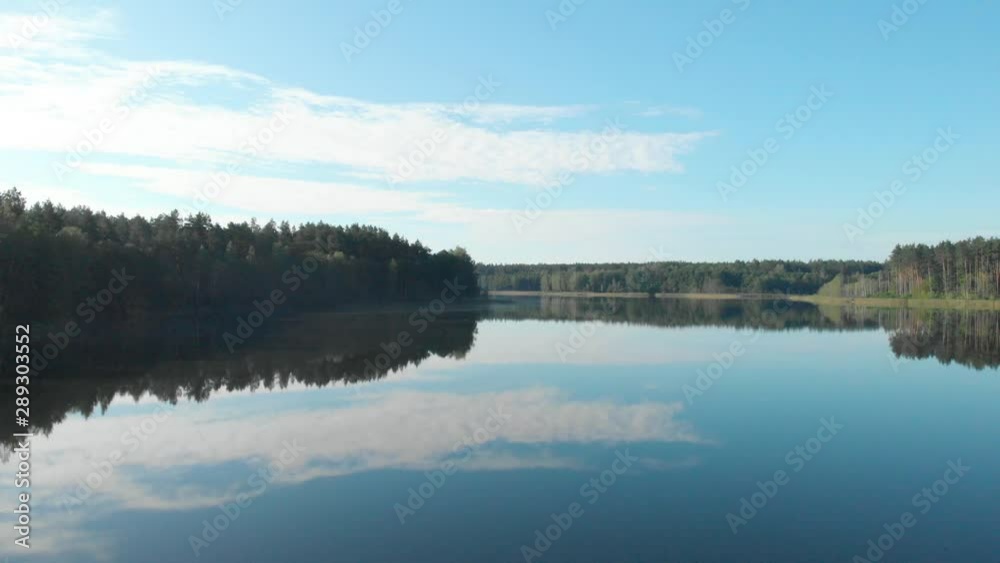 Slow aerial flying over a calm, empty lake reflecting the blue sky and clouds in the Lithuanian countryside