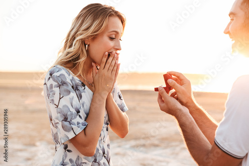 Photo of smiling man making proposal to his amazed woman with ring while walking on sunny beach photo