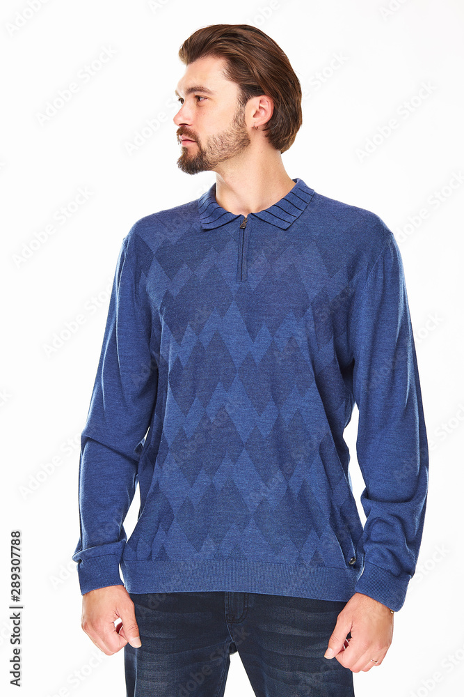 Fototapeta premium man with beard and dark hair stands on a white background in blue sweater with zip and collar and dark jeans, he has a gold watch on his arm and looks on the left direction