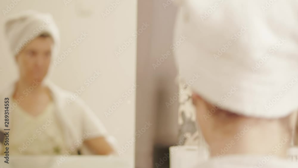 Woman with wrapped hair in white towel after washing head looking to mirror, taking off earrings in hairdressing beauty saloon, focus on face in glass, then to hands and nape. Lady before haircut Woman with wrapped hair in white towel after washing head looking to mirror, taking off earrings in hairdressing beauty saloon, focus on face in glass, then to hands and nape. Lady before haircut