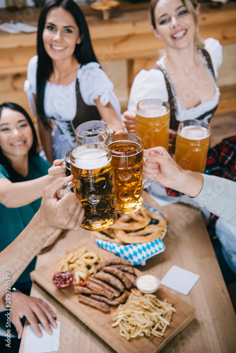 selective focus of attractive waitresses in traditional german costumes clinking mugs of beer with multicultural friends