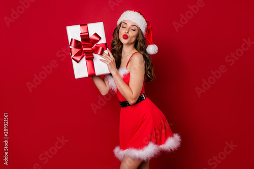 Pretty lady holding big giftbox in hands sending air kiss to handsome santa wear fluffy mini dress isolated red background