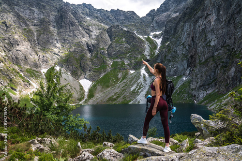 Young brunette girl in red sportswear with backpack standing near the mountain lake and points a hand into the distance