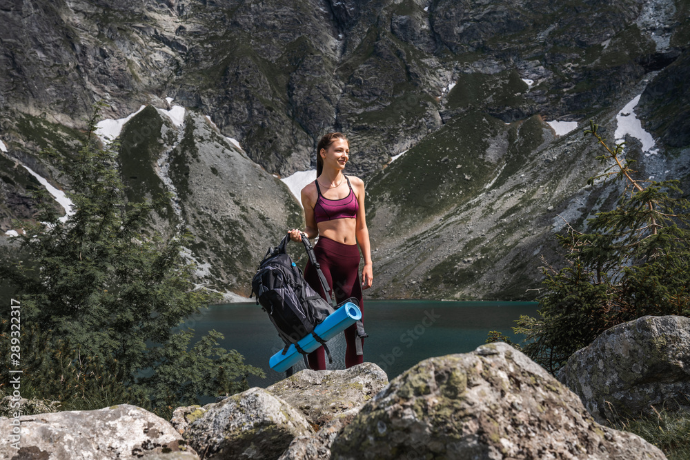 Young brunette girl traveler in red sportswear with backpack in hand standing on the stones, amazing mountine lake on the background, active and healthy lifestyle concept