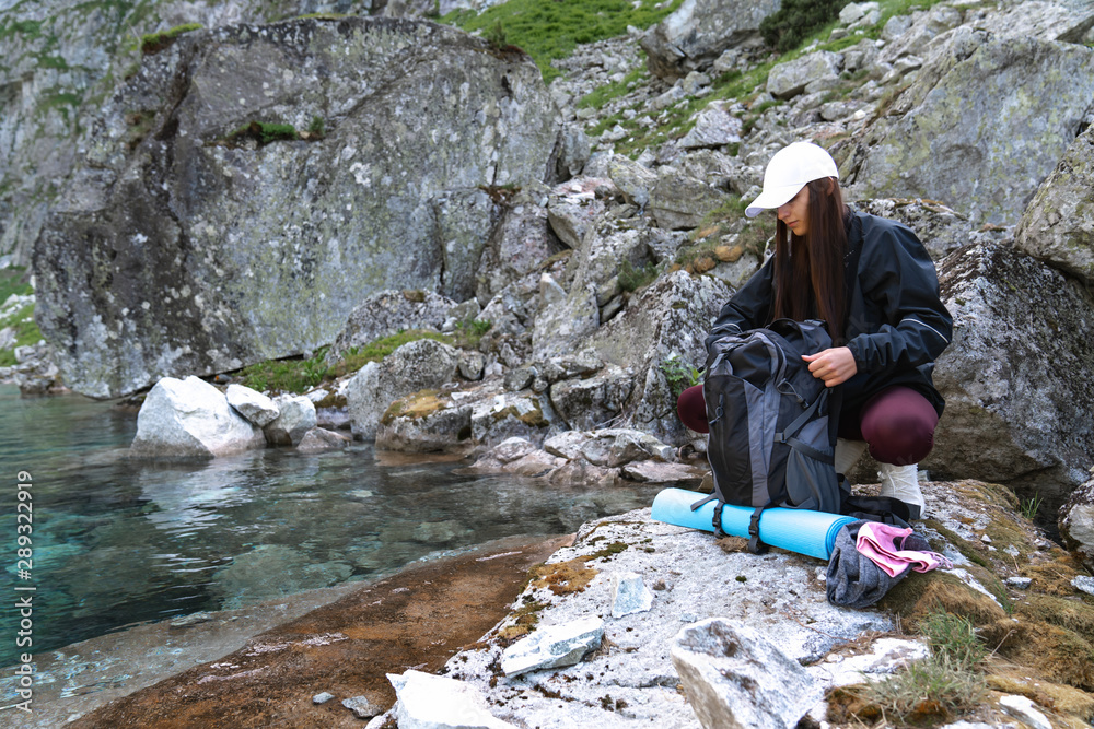 Girl traveler in white hat and black jacket sitting near the mountain lake and opens the backpack