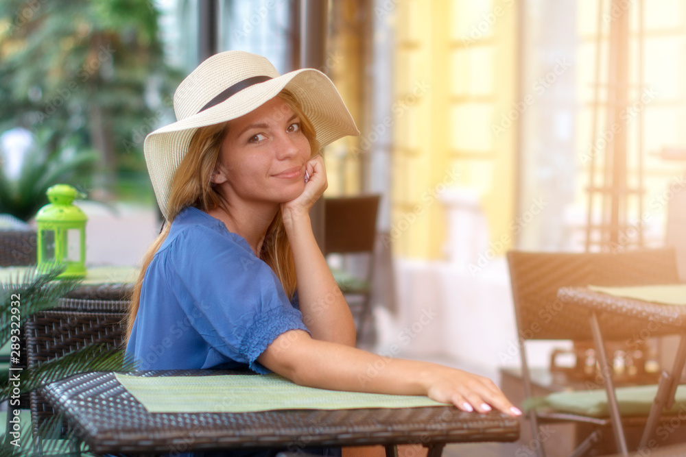 A beautiful slender girl with a sweet smile and a light feminine hat with a brim in a street cafe on the street.