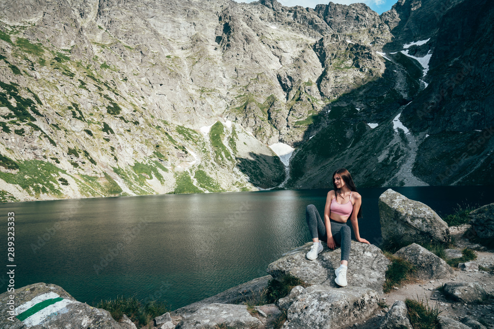 Young beautiful sporty girl in grey pants sitting on the big rock and posing on the background of mountain lake, active lifestyle concept Young beautiful sporty girl in grey pants sitting on the big rock and posing on the background of mountain lake, active lifestyle concept
