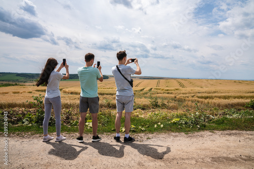 Three friends travelers in casual clothes standing back to the camera and making phone photo of beautiful panorama