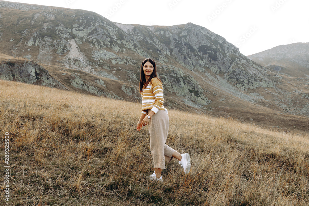 Stylish girl in rustic dress smiling and waving hair in sunny meadow in mountains. Portrait of happy boho woman in countryside at sunset, positive vibes, rural simple life. Atmospheric image