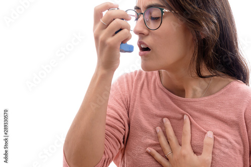 Young caucasian brunette girl with pink shirt suffers from asthma inhaling with an inhaler. isolated in white background.