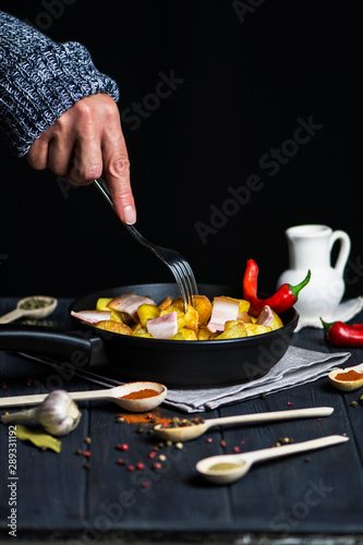 hand holds a fork with fried potatoes and bacon meat over a pan on a dark background