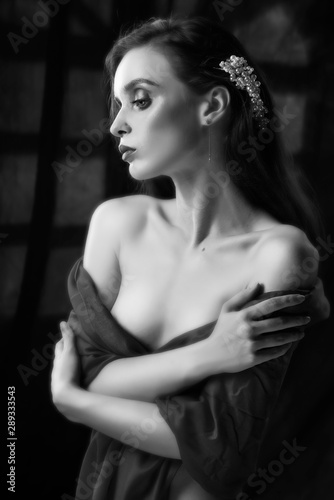 Beautiful girl with fashionable makeup, wearing a silk scarf thrown over her shoulders that covers her naked breast, sensually poses on the background of the shadow from the window. Black and white
