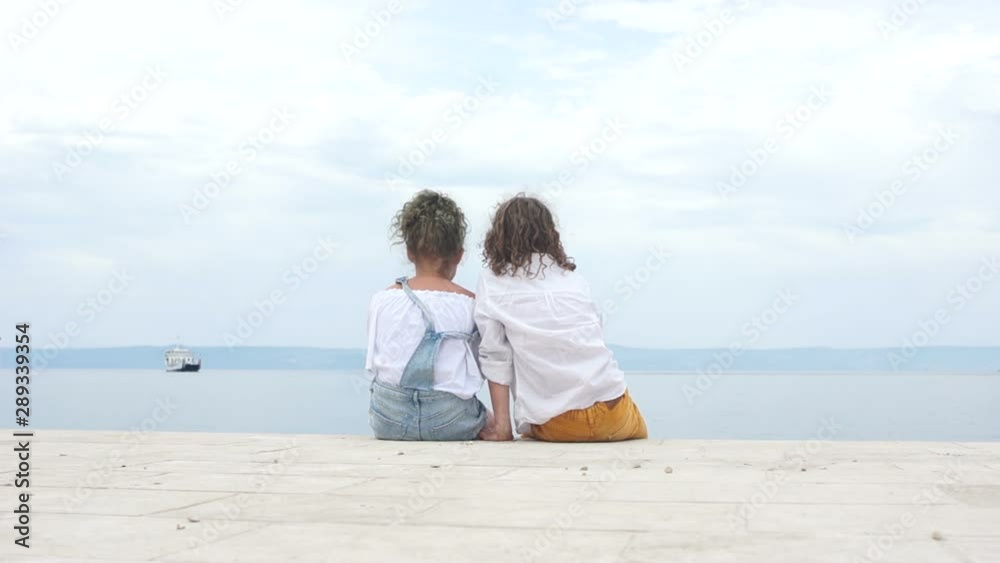 Boy and a girl schoolchildren are sitting next to the seashore and looking into the distance at a sailing ship. Travel concept, summer vacation Boy and a girl schoolchildren are sitting next to the seashore and looking into the distance at a sailing ship. Travel concept, summer vacation