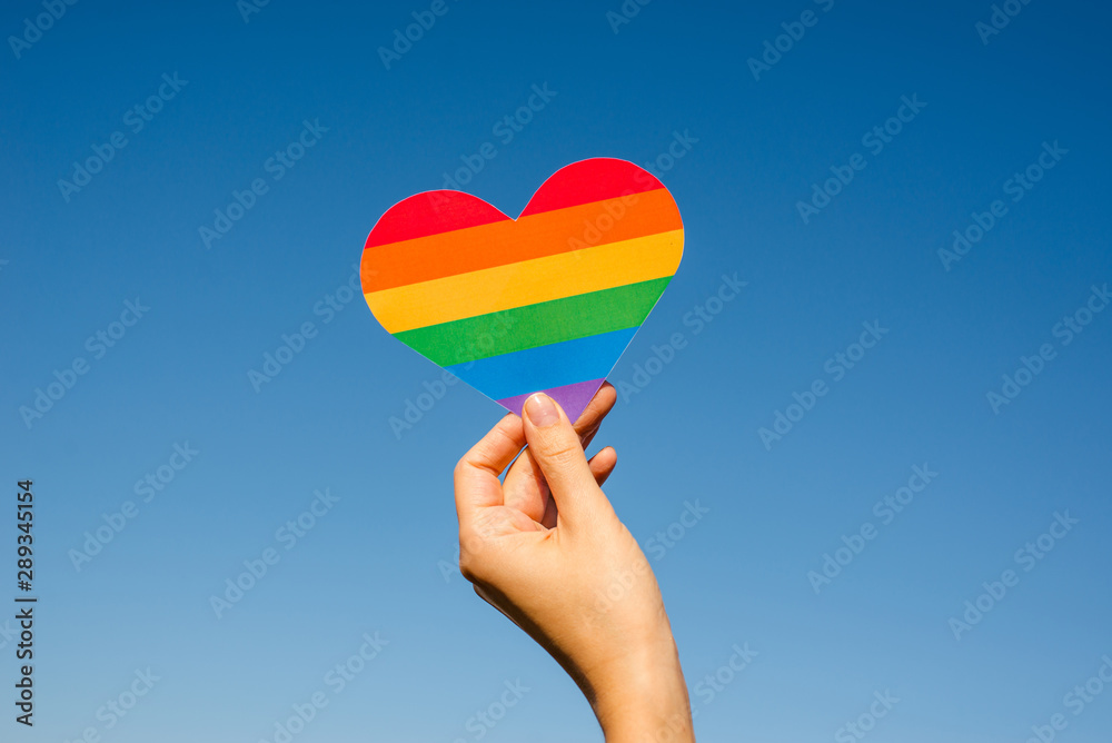 Woman holds in hands a heart in the colors of the rainbow. Young beautiful girl. LGBT history month. Pride Month. Lesbian Gay Bisexual Transgender. LGBT flag. Love, human rights, tolerance. LGBTQ+ - obrazy, fototapety, plakaty 