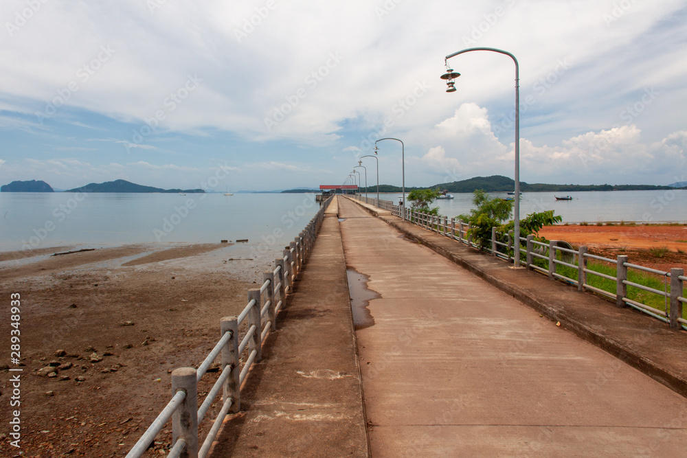Long pier at Koh Phangan island, Thailand, during low tide with clouds, boats, and mountains on the background.