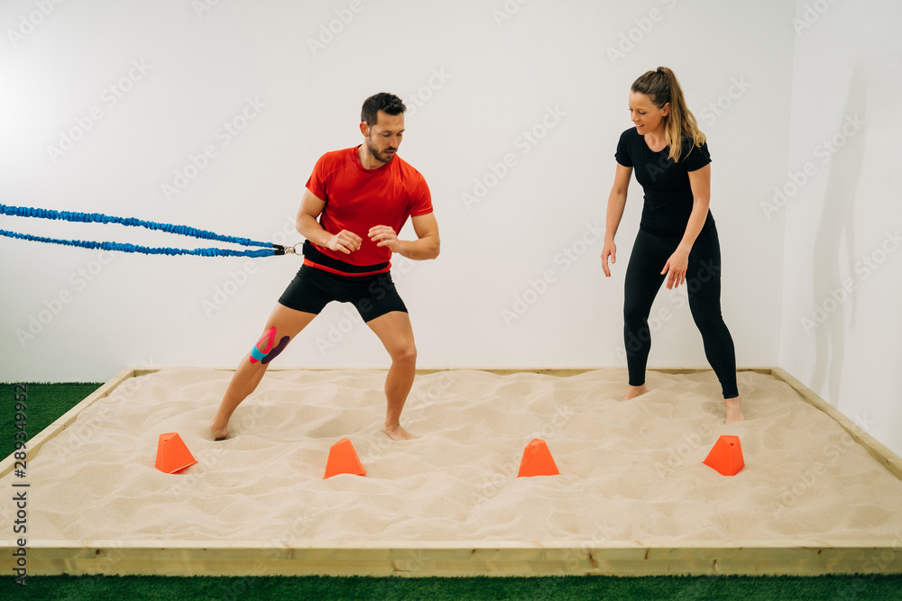 Man does stretching exercises on sand with a blue support at the waist, assisted by a physiotherapist girl, a white wall in the background. Concept of muscle health and relaxation.