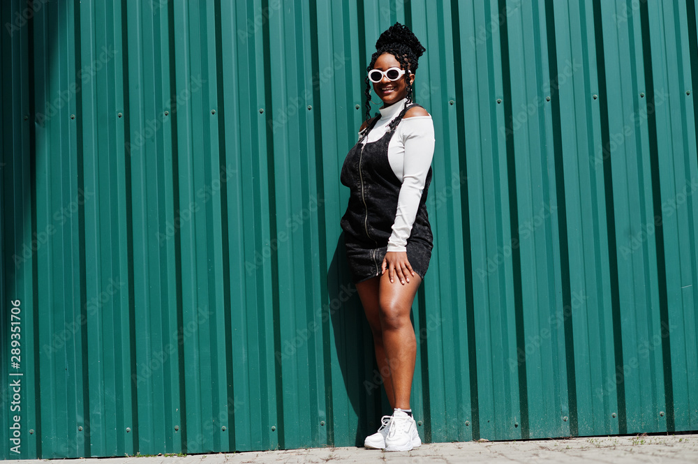 African woman in denim overalls skirt, white sunglasses posed against green steel wall. African woman in denim overalls skirt, white sunglasses posed against green steel wall.