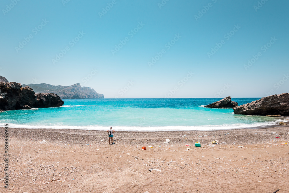 Olympos, Karpathos/ Greece - 07 09 2019: volunteer woman starting beach cleanup and walking on a beautiful beach full of plastic rubbish litter in background is the blue Aegean Sea holiday destination