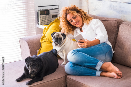 Cheerful woman at home sit down on the sofa with her two best friends dog pug near her - real lifestyle people with animals