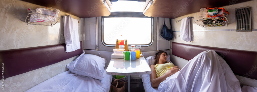 Panorama of the interior of a reserved seat train car, a girl sleeping on one of the lower shelves - obrazy, fototapety, plakaty 