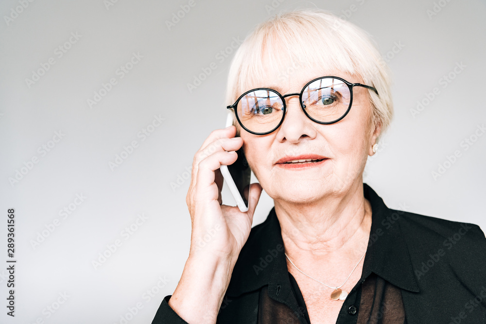 senior businesswoman in black outfit and glasses talking on smartphone isolated on grey