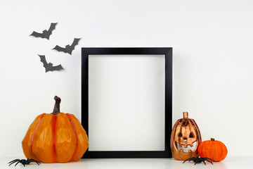Mock up black frame with Jack o Lantern and pumpkin decor on a shelf or desk. Halloween concept. Portrait frame against a white wall with bats.
