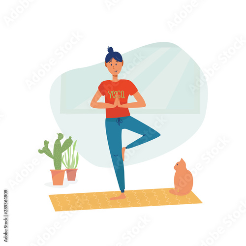 Girl in t shirt with sign Yoga in tree pose practice asana balance exercise at home with smile, cat sitting behind. Healthy, home training and sport activity concept. 