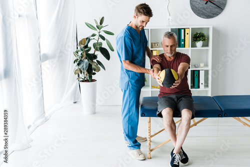 handsome doctor standing near mature man working out with ball photo