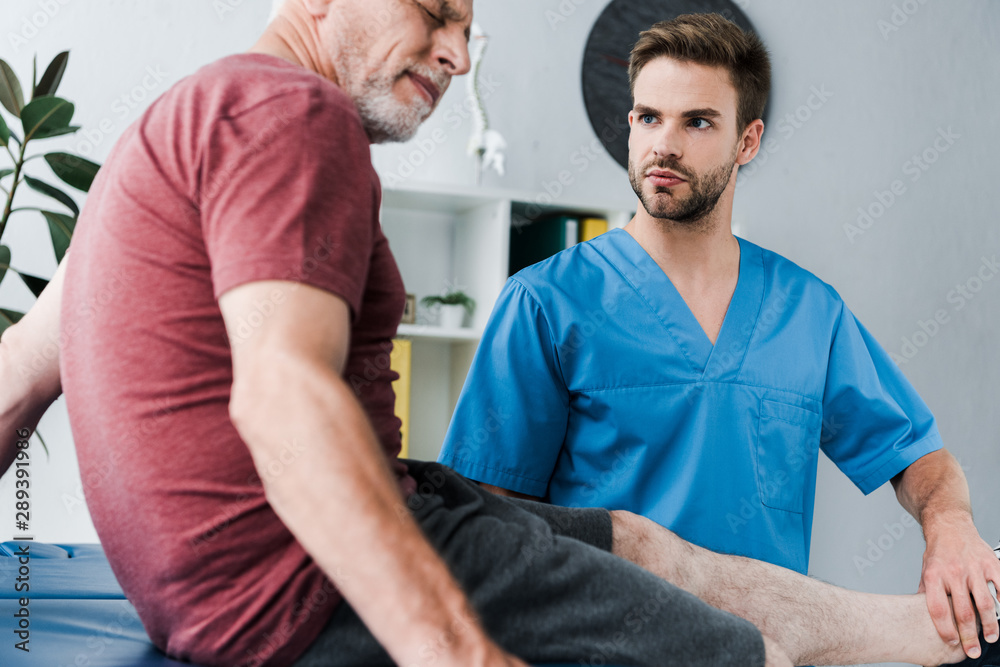 Obraz premium selective focus of handsome bearded doctor holding leg on mature patient with pain