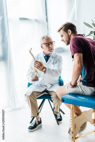 bearded doctor in glasses holding spine model and looking at patient