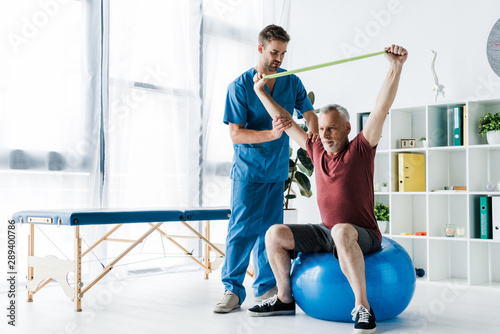 doctor standing near middle aged man exercising with resistance band while sitting on fitness ball