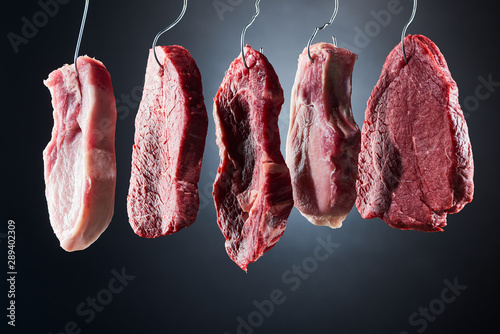 Tablou pe pânză assorted raw meat on metal hooks on dark black background