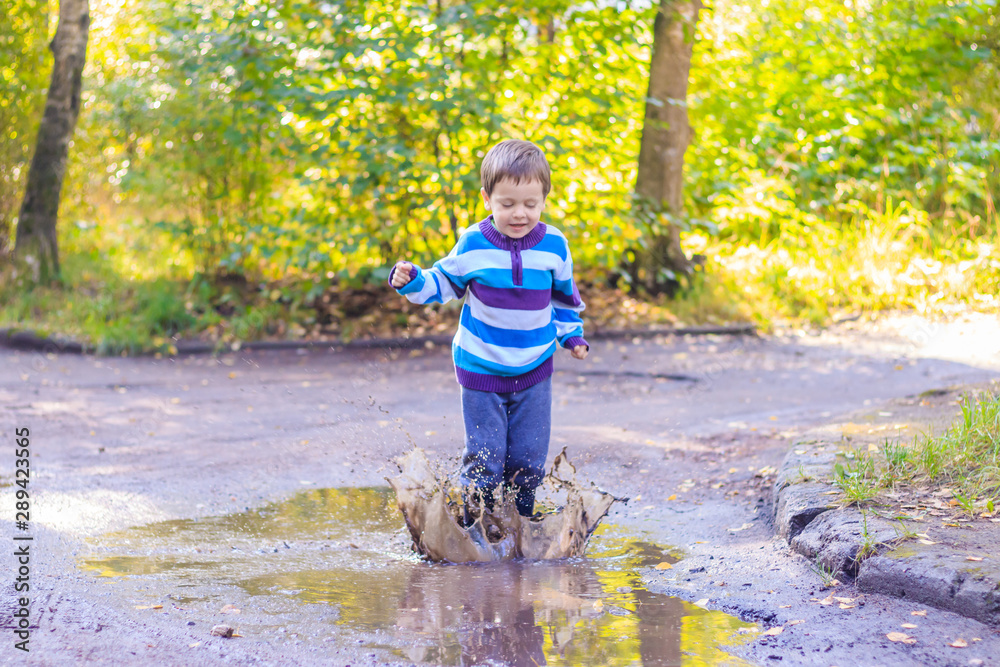 A little boy is jumping in a puddle. A boy in rubber boots. Happy childhood. Puddles after the rain. Warm summer evening. A little boy is jumping in a puddle. A boy in rubber boots. Happy childhood. Puddles after the rain. Warm summer evening.