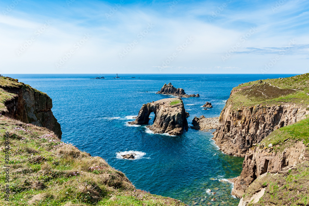Dry grassy cliffs overlooking Enys Dodnan Arch on a partially sunny day in Land’s End, UK.