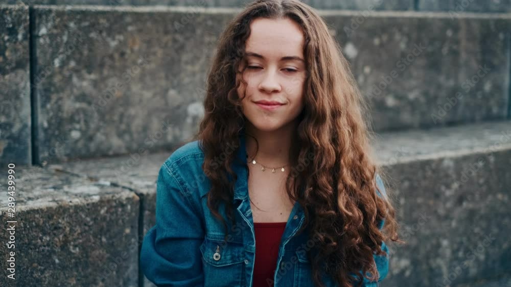 Close up of the cheerful Caucasian teenage girl looking at the side, then turning face to the camera and smiling while resting outdoors on the stoned steps. Portrait. Close up of the cheerful Caucasian teenage girl looking at the side, then turning face to the camera and smiling while resting outdoors on the stoned steps. Portrait.