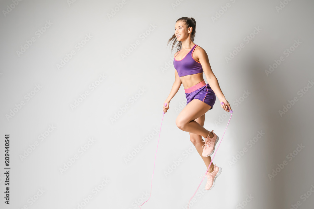 Sporty young woman jumping rope against light background