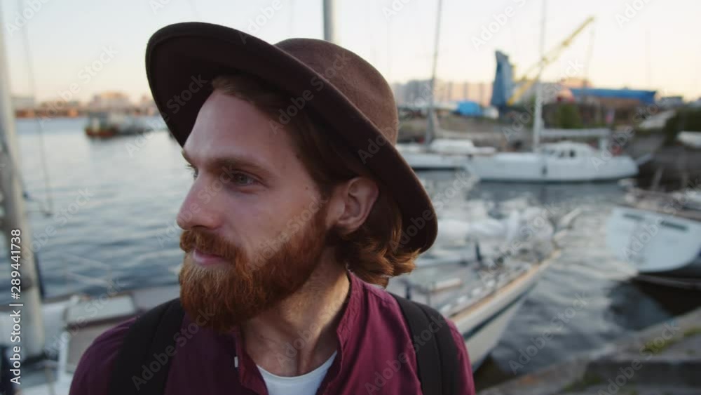 Close up of Caucasian man with red beard wearing street wear and hat standing on embankment, smiling while looking around Close up of Caucasian man with red beard wearing street wear and hat standing on embankment, smiling while looking around